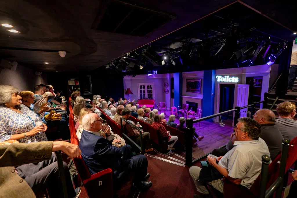 The audience seated around a small purple-lit stage at Jermyn Street Theatre