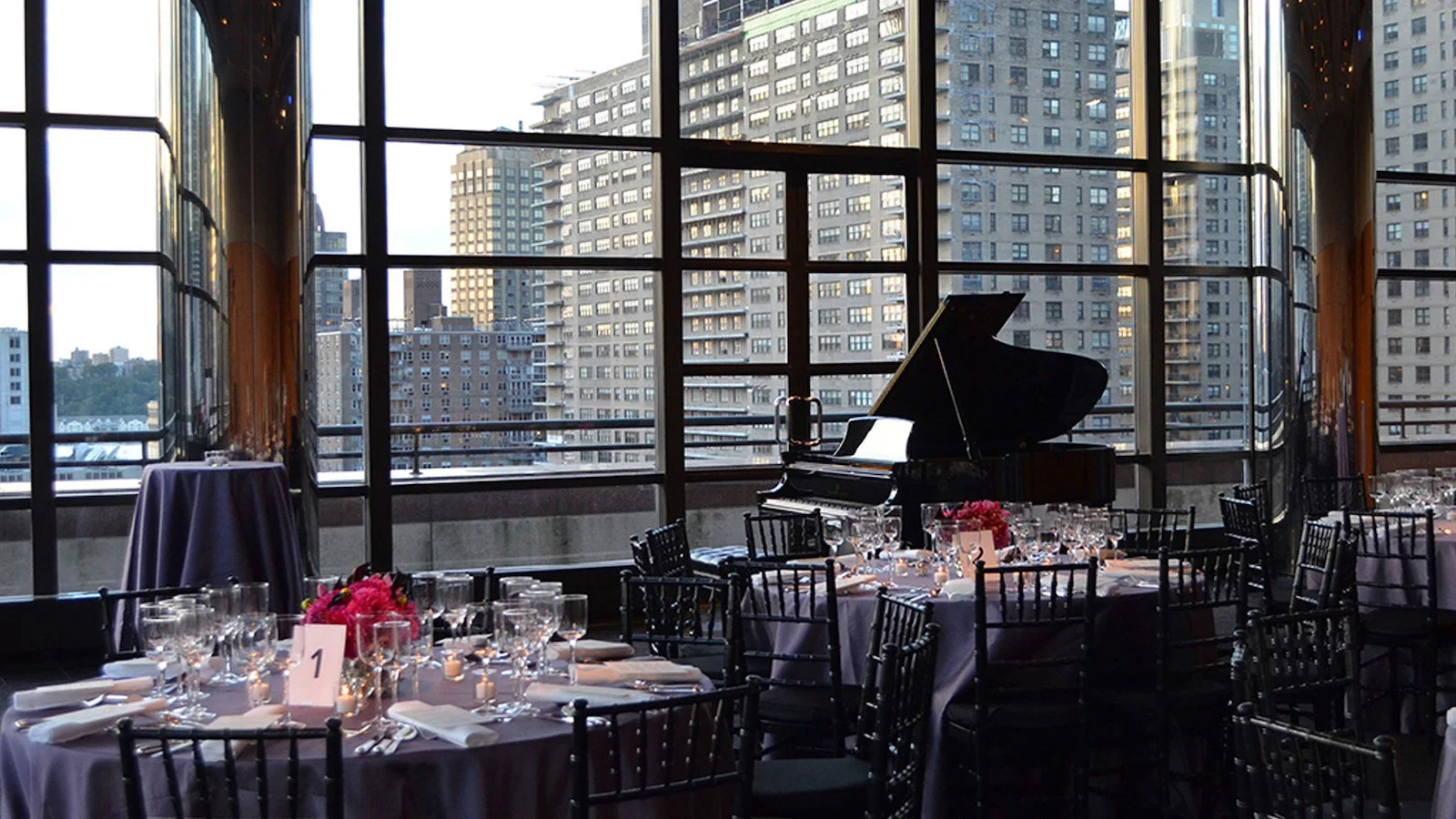 A piano and tables beside tall city-facing windows at Stanley H. Kaplan Penthouse