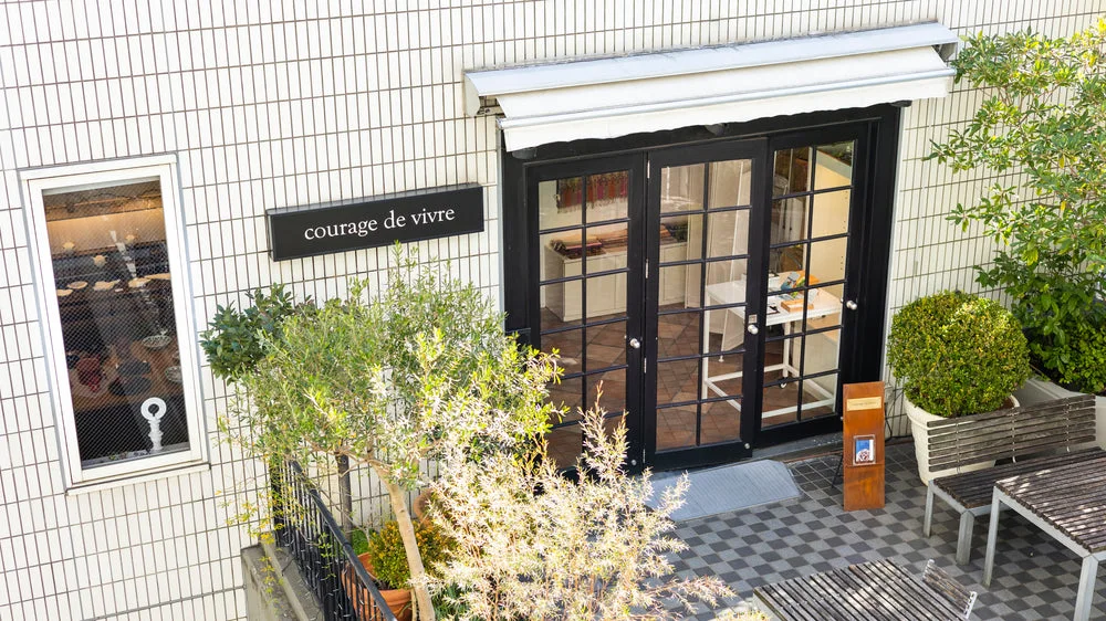 The gallery's glass entrance framed by small plants at Courage de Vivre
