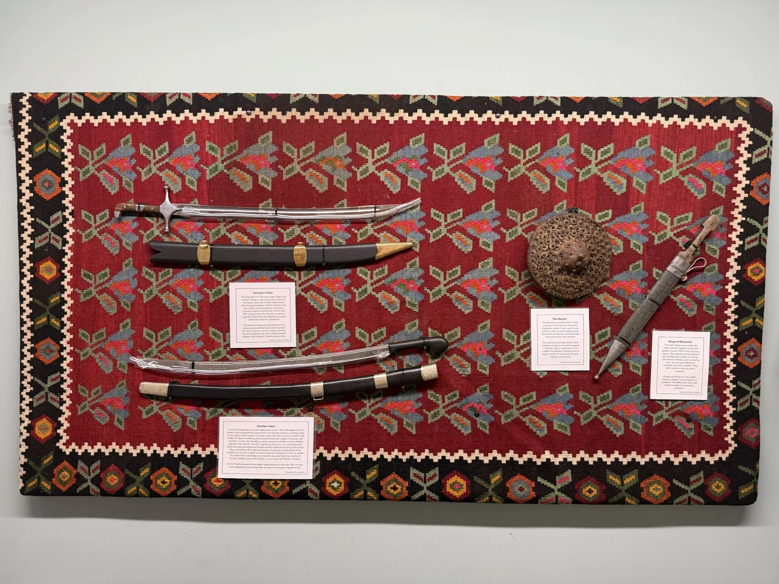 Swords and a shield displayed on red fabric in the Armenian collection at the Martial Arts History Museum in Glendale, photographed in 2025